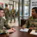an image of two Soldiers sitting at a table in a military police station underneath a sign that says Military Police Investigations CID. One of the Soldiers is an MP wearing an CID investigators badge. Military Law Enforcement Investigations: What should I expect?